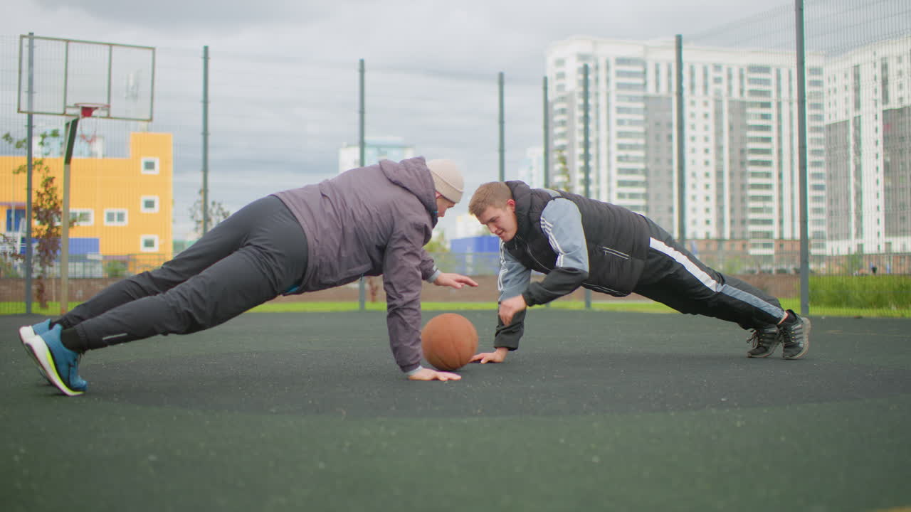 Two white men doing partner pushups on outdoor basketball court, urban highrise backdrop, basketball placed between palms, hooded jackets and sneakers, cooperative rivalry with trainer and trainee