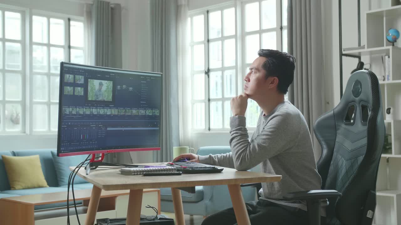 Asian Video Editor Man In Long Sleeved T-Shirt And Black Pants Thinking While Using Desktop Computer For Editing Video At Home.