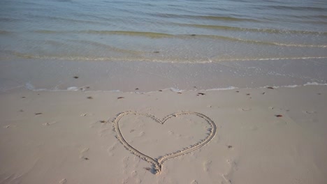 wide angle static shot of heart with an arrow through it on a beach with water lapping on the sand