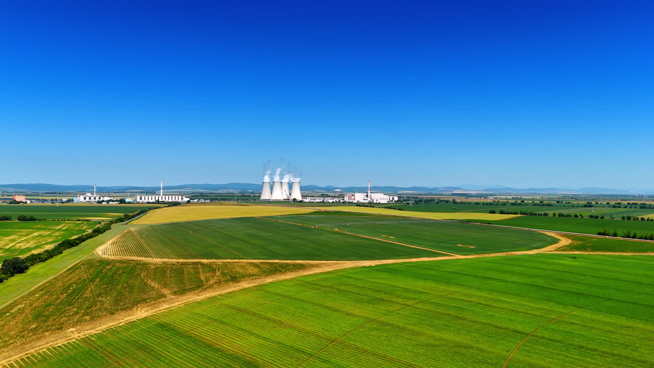 Plant by green fields, blue sky. Smoke rises from cooling towers of a power plant set against a backdrop of lush green fields under a clear blue sky