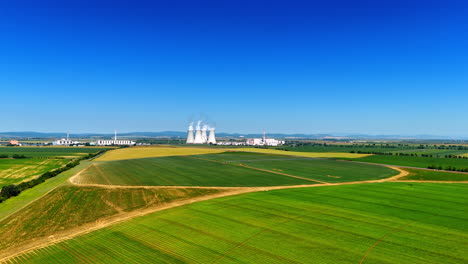 plant by green fields, blue sky. smoke rises from cooling towers of a power plant set against a backdrop of lush green fields under a clear blue sky
