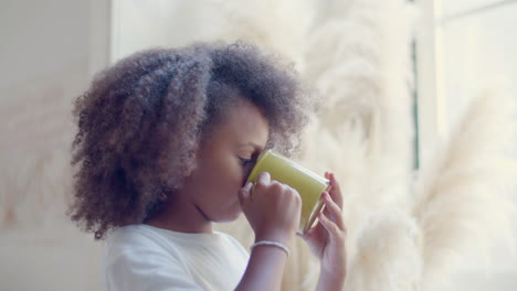 cute black girl standing by the window, drinking tea, then turning to camera and smiling