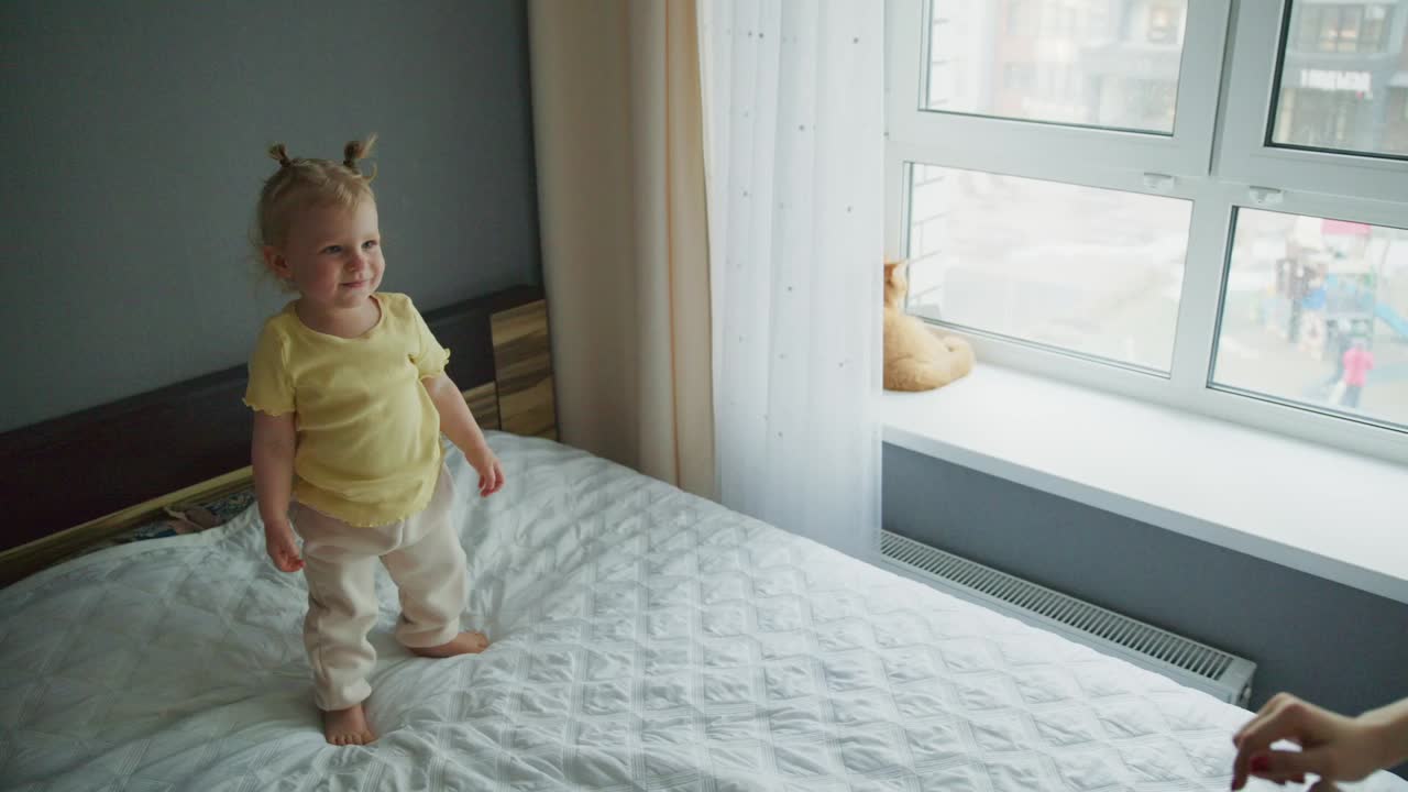 Süßes Kleinkindmädchen steht auf dem Bett und lächelt einen Elternteil an.