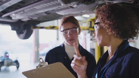 Nachhilfelehrerin-Mit-Studentin,-Die-Im-Kfz-Mechaniker-Kurs-Auf-Einer-Hydraulischen-Rampe-Unter-Das-Auto-Schaut