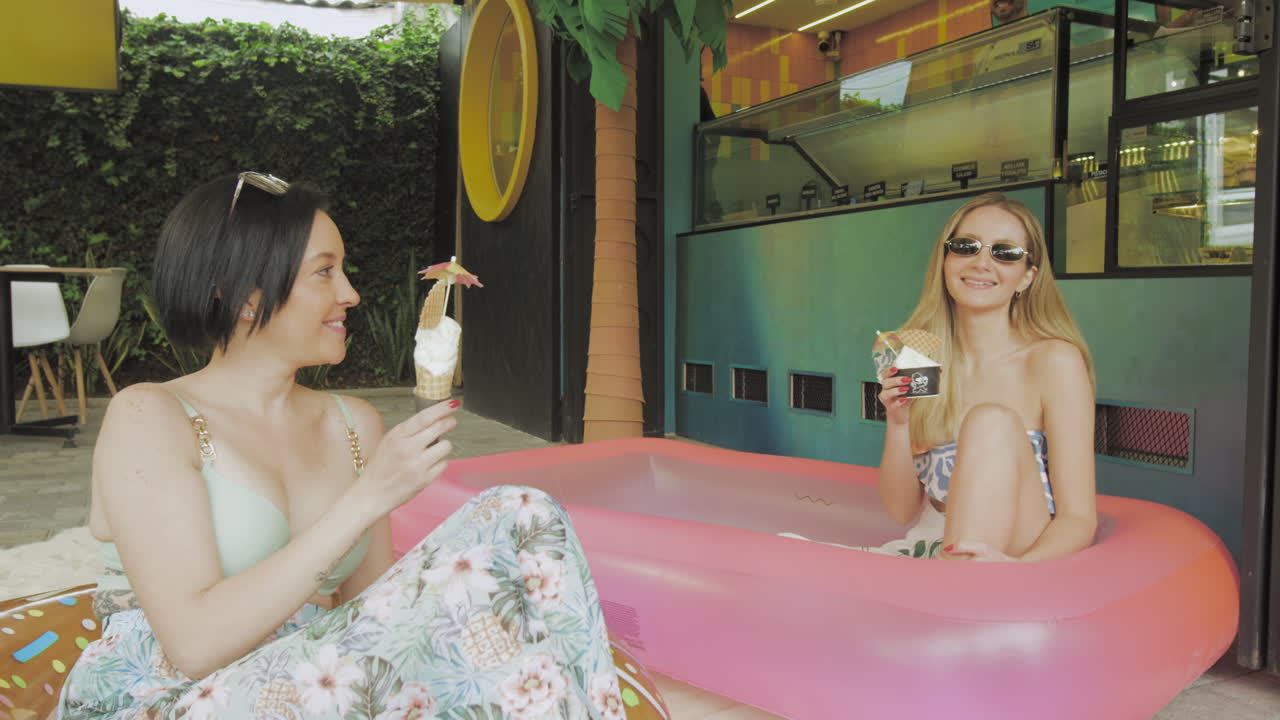 Two young women enjoy ice cream in a relaxed atmosphere next to an inflatable pool in front of a contemporary ice cream parlor