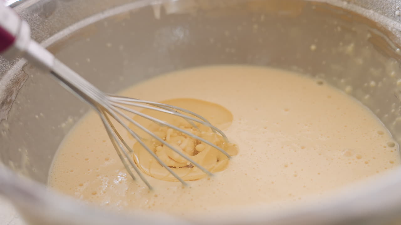 Close up of oil being poured into flour mix as caterer stirs with whisk in stainless bowl, showing preparation of homemade batter during cooking process in bright kitchen setting
