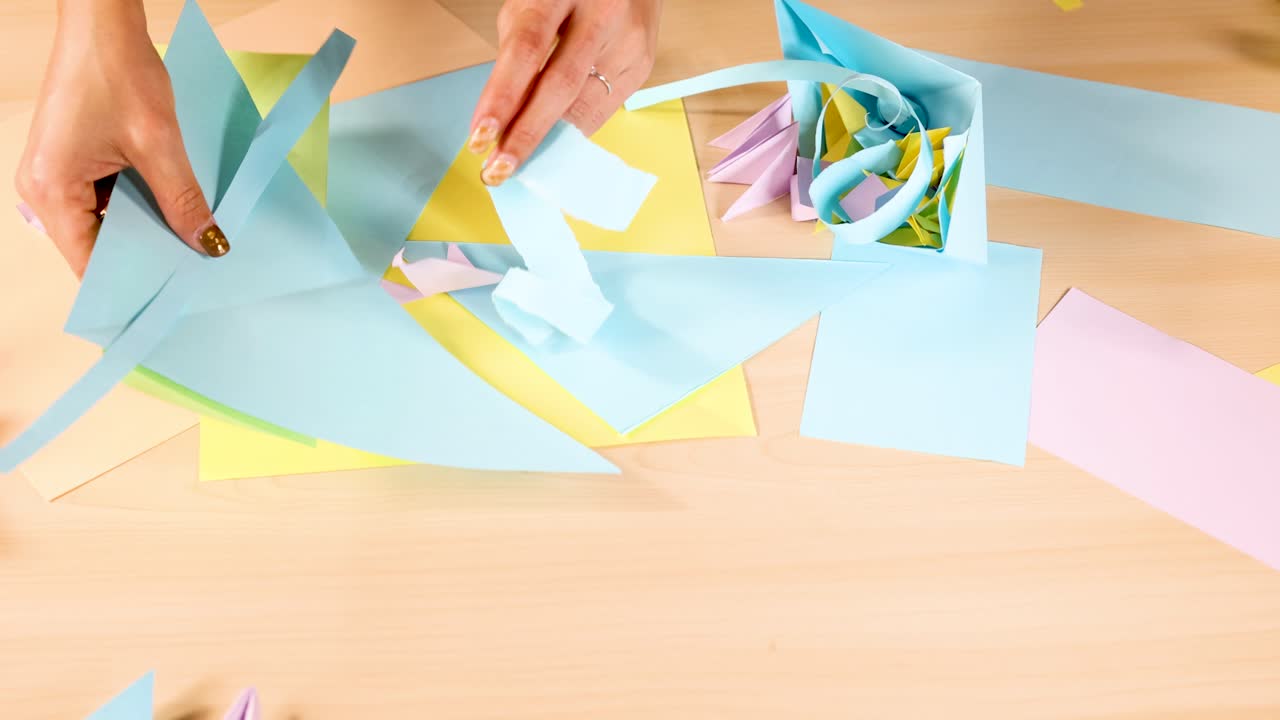 Person creates pastel origami flowers using colored paper on wooden table, top-down view, bright lighting
