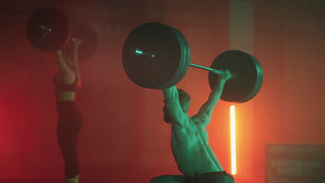 a man and a woman in a colored neon red and blue light in slow motion lift heavy barbells over their heads. strength and power training with heavy weight. the concept of athletic and strong couples