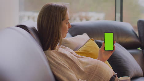 Pregnant-Woman-With-Prosthetic-Arm-On-Sofa-At-Home-With-Hot-Drink-Holding-Phone-With-Green-Screen