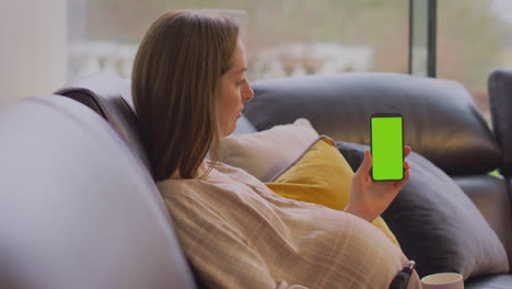 mujer embarazada con brazo protésico en el sofá en casa con bebida caliente sosteniendo el teléfono con pantalla verde