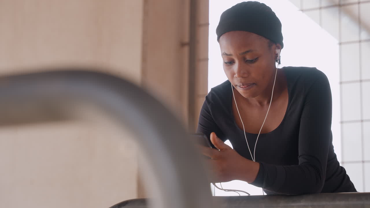 Donna nera su un balcone che sistema gli auricolari mentre controlla il telefono, luce solare delicata, architettura minimalista in cemento, collana di perline, umore tranquillo e riflessivo, top nero casual, ritratto spontaneo.