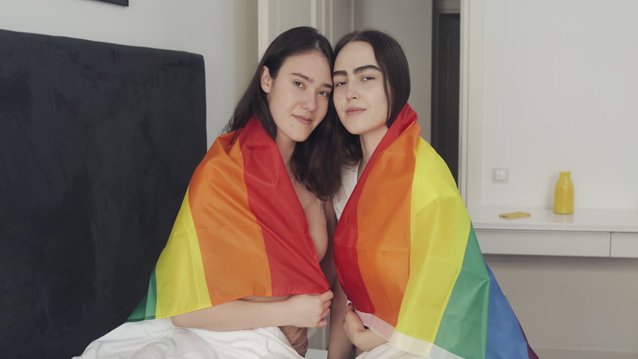 Two women wrapped in rainbow flag in bed