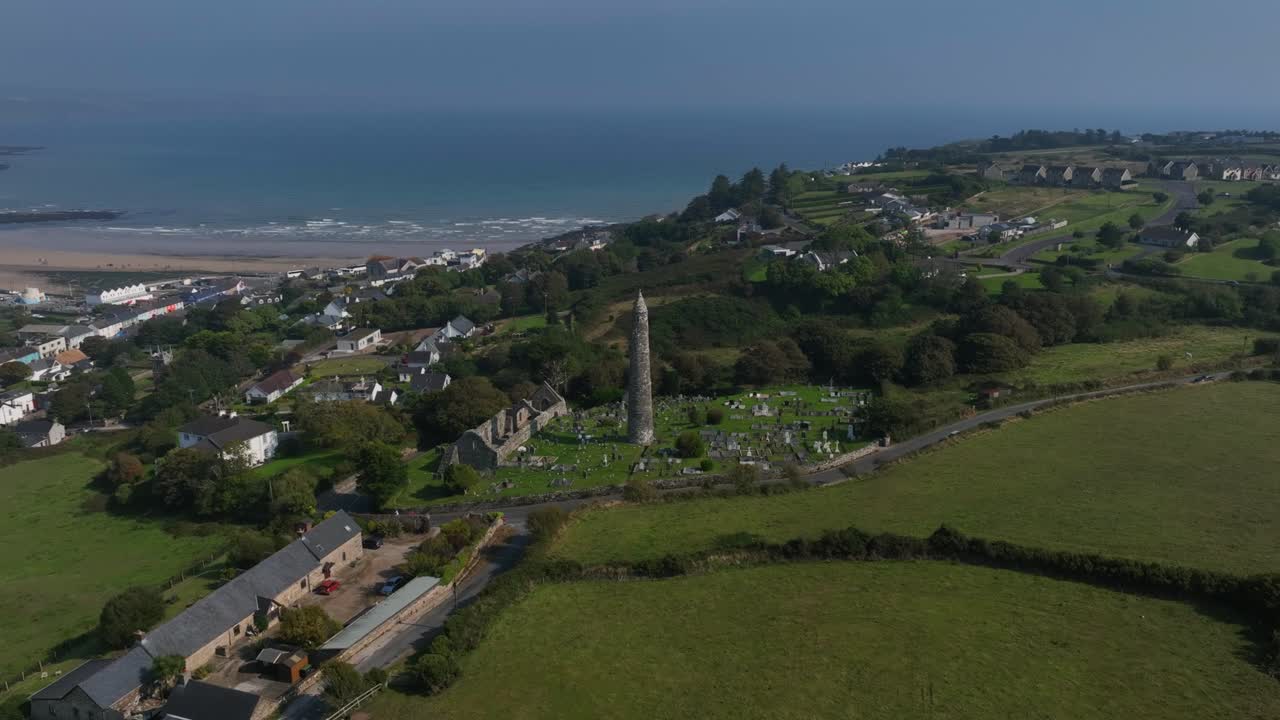 St. Declan's Monastery, Ardmore, County Waterford, Ireland, September 2024. Drone orbits counter clockwise pulling backward around the Round Tower as waves crash and roll towards the distant beach.