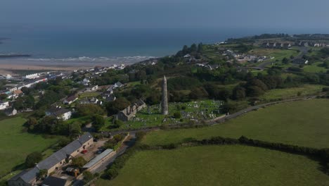 st. declan's monastery, ardmore, county waterford, ireland, september 2024. drone orbits counter clockwise pulling backward around the round tower as waves crash and roll towards the distant beach.