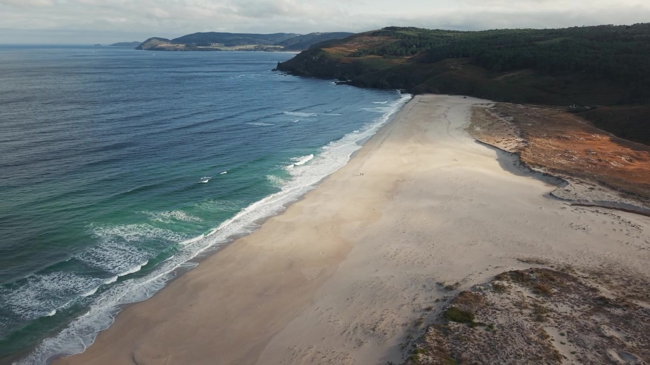 Drone pulling back above Praia do Rostro revealing a vast Atlantic beach with small silhouettes walking along the shore.