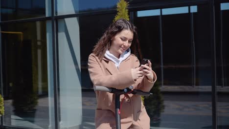 a young student girl in a sweater, jeans and a coat with an electric scooter stands near a stylish modern shopping center building and uses a smartphone. front view