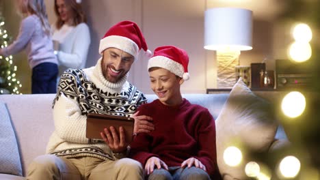 Portrait-Of-Joyful-Dad-With-Teen-Boy-Sitting-In-Cozy-Room-Typing-On-Tablet-Buying-Xmas-Gifts-On-Internet