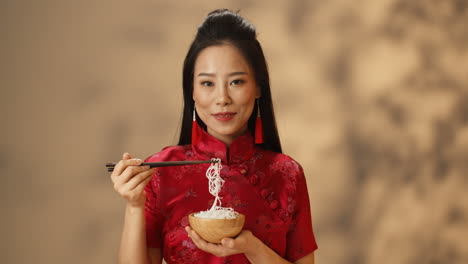 retrato de una joven asiática alegre vestida con ropa tradicional roja comiendo fideos y sonriendo a la cámara