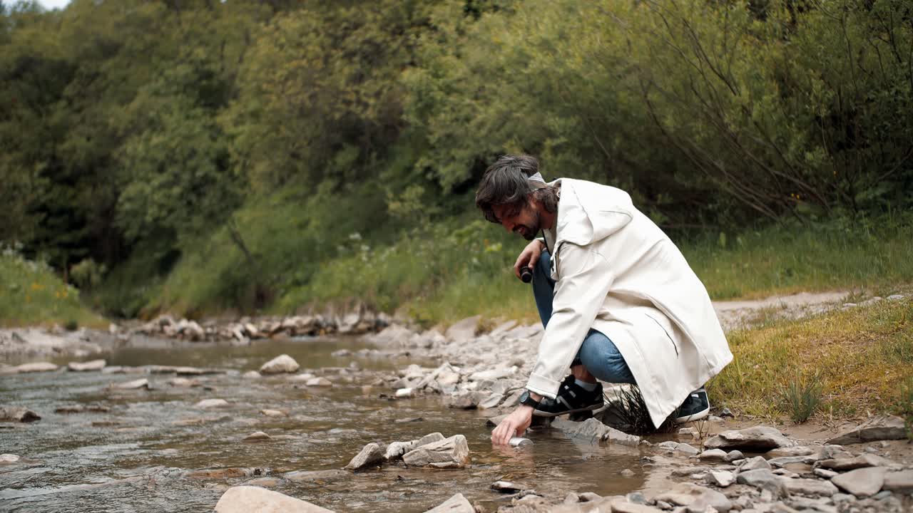 un tipo con una chaqueta blanca de senderismo extrae agua en un termo de un río de montaña mientras camina en el bosque