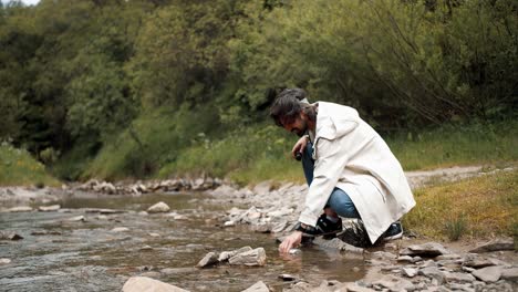 A-guy-in-a-white-hiking-jacket-draws-water-into-a-thermos-from-a-mountain-river-while-hiking-in-the-forest