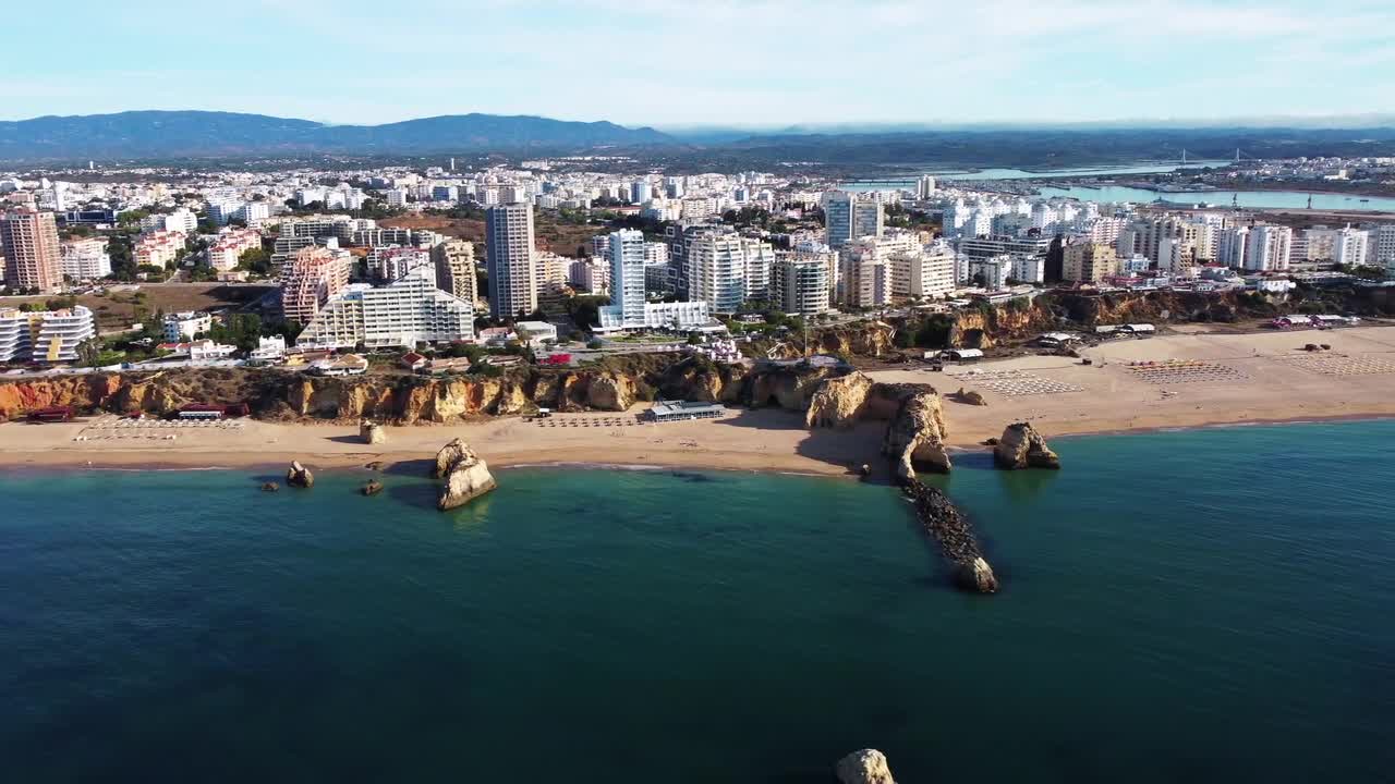 Aerial view of modern city buildings along sandy beach with rocky cliffs and calm blue sea in Faro, Portugal. Clear sky, coastal charm, and urban contrast create a vibrant scenic atmosphere.