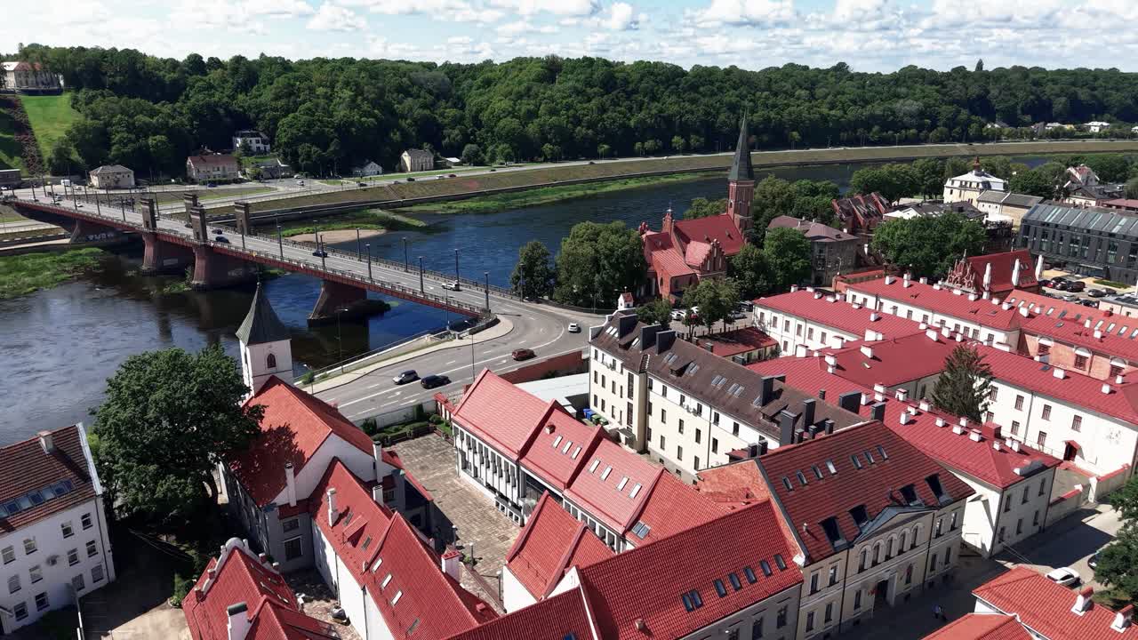 Aerial view of a historic European town with red rooftops, a river, and a bridge, surrounded by lush greenery and architectural landmarks