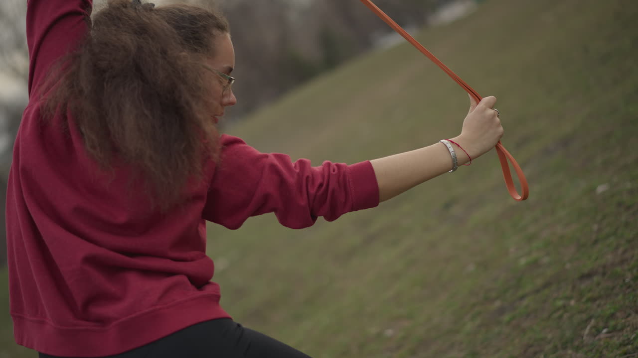 Focused White Woman Rotating Orange Band Across Back In Park, Dancer Warming Up For Rehearsal With Controlled Rotation And Flexibility Emphasis, Cloudy Sky And Quiet Field Setting
