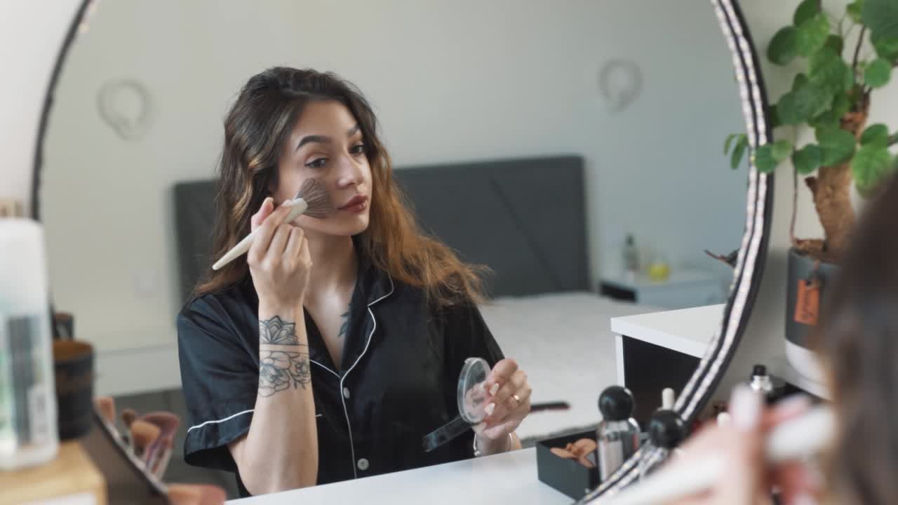 retrato de una joven hermosa mujer en pijama sentada frente al espejo aplica maquillaje con un pincel, aplicando polvo facial