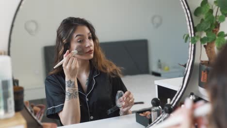 portrait of a young beautiful woman in pajamas sitting in front of the mirror applies makeup with a brush, applying face powder