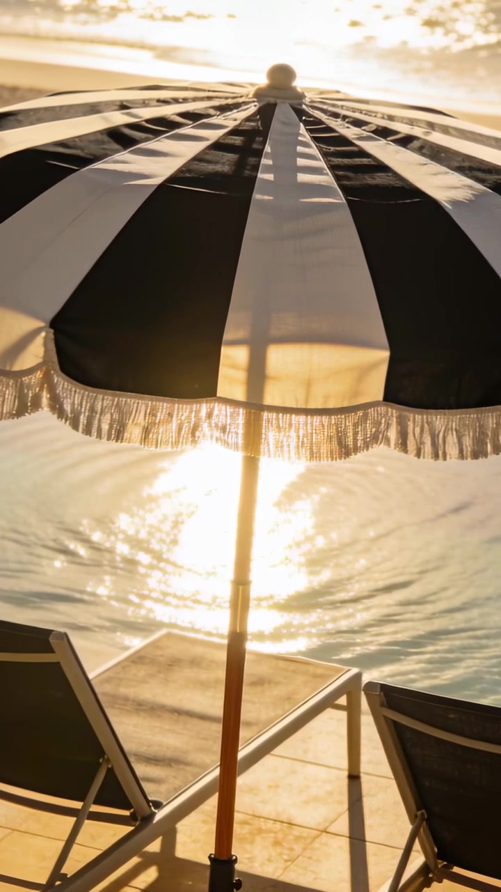 Vidéo verticale : Le soleil bas projette l'ombre d'un parasol rayé et fait scintiller l'eau sur les transats de la terrasse.