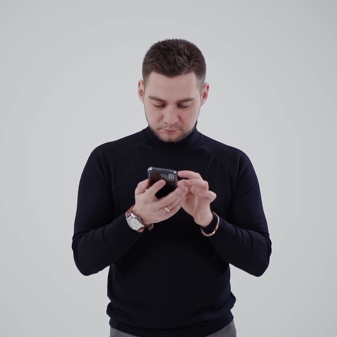 Handsome man with a cell phone isolated in studio. Portrait of a young man in black sweater and grey trousers looking in his phone.