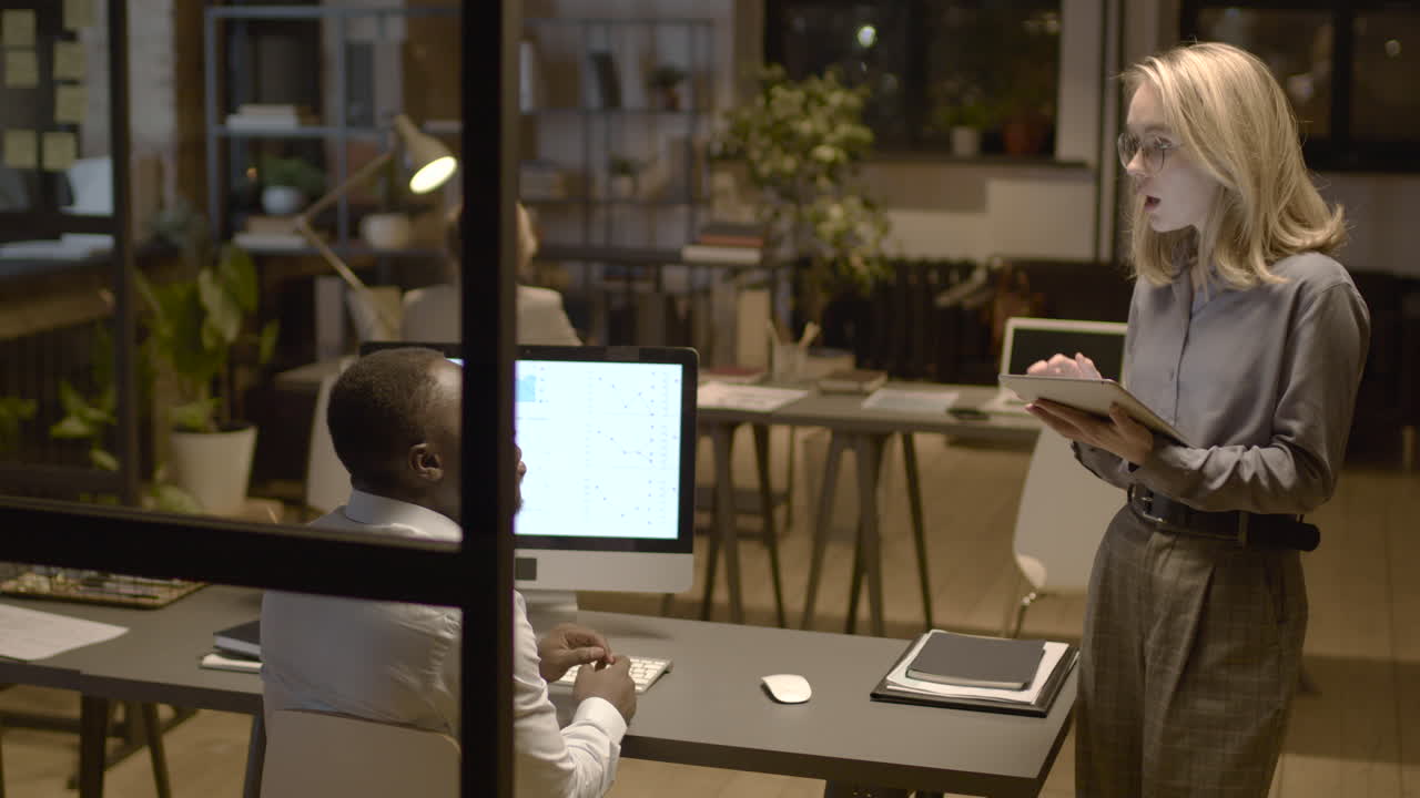 Rear View Of American Man Sitting At Desk While Talking With Female Coworker Who Is Holding A Tablet In The Office