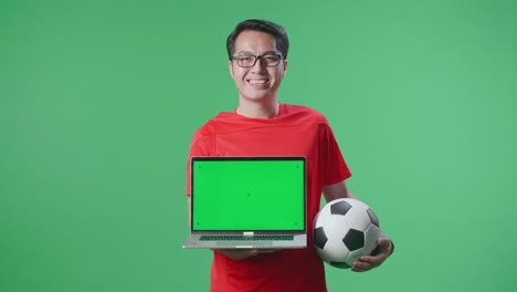 hombre asiático con una pelota que muestra una maqueta de portátil de pantalla verde y sacudiendo la cabeza mientras anima el fútbol en el fondo de la pantalla verde