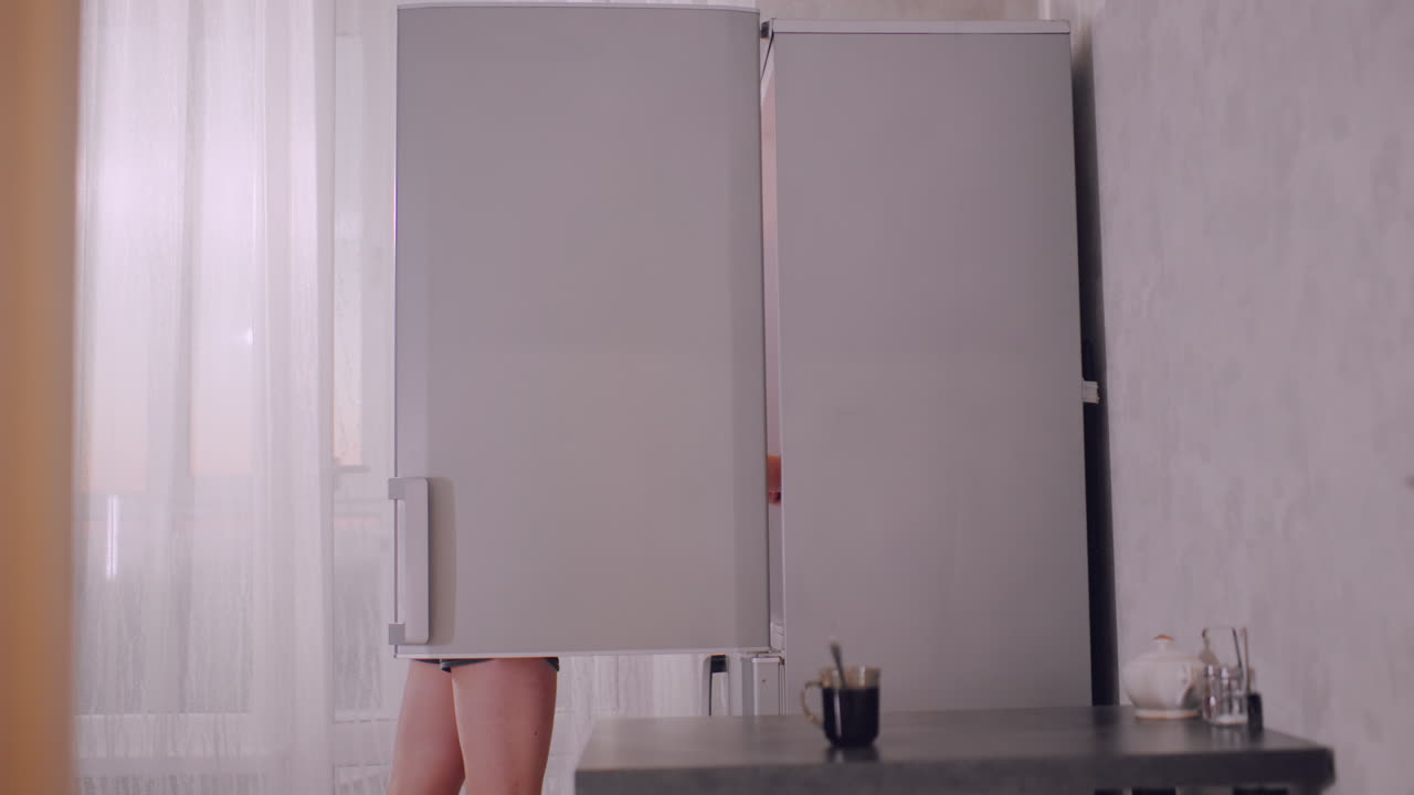 Casual woman in shorts opens fridge in bright kitchen, preparing breakfast by taking out two plates of bread while cup sits on table, morning sunlight softly filtering through sheer curtains