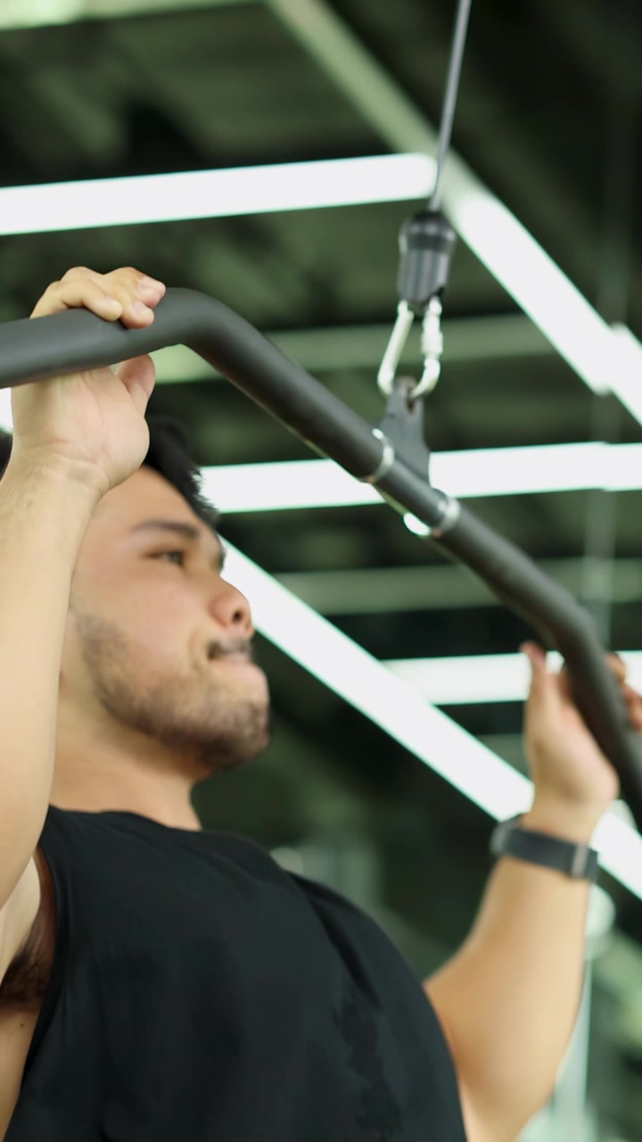 Focused young Asian man doing lat pulldown workout on cable machine in bright modern gym.