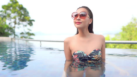 portrait of an asian woman in big red sunglasses and swimsuit inside the swimming pool water turning her sight, slow-motion handheld