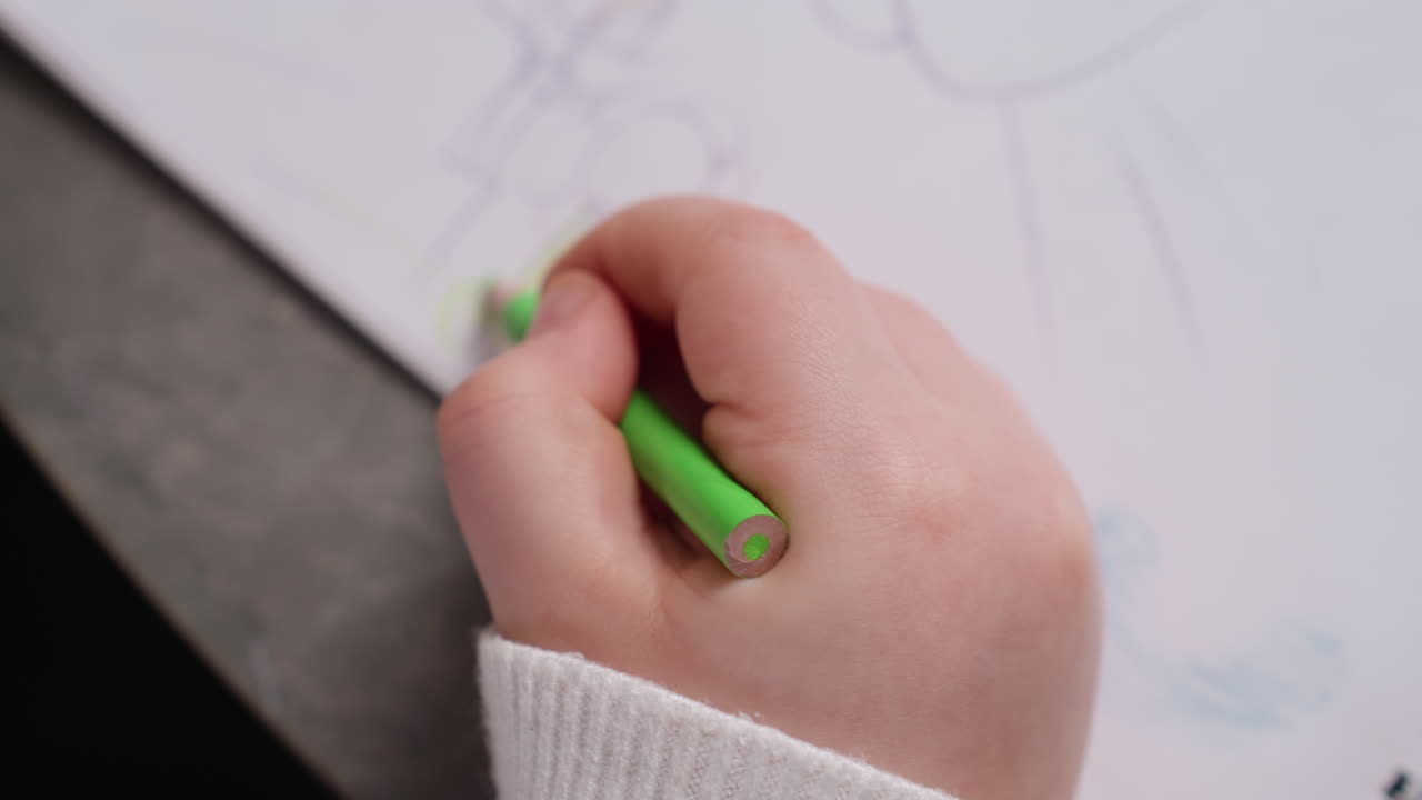 Close up of student hand using green pencil to draw simple sketch of tree and house on white notebook paper, symbolizing imagination, creativity, early learning, childhood art