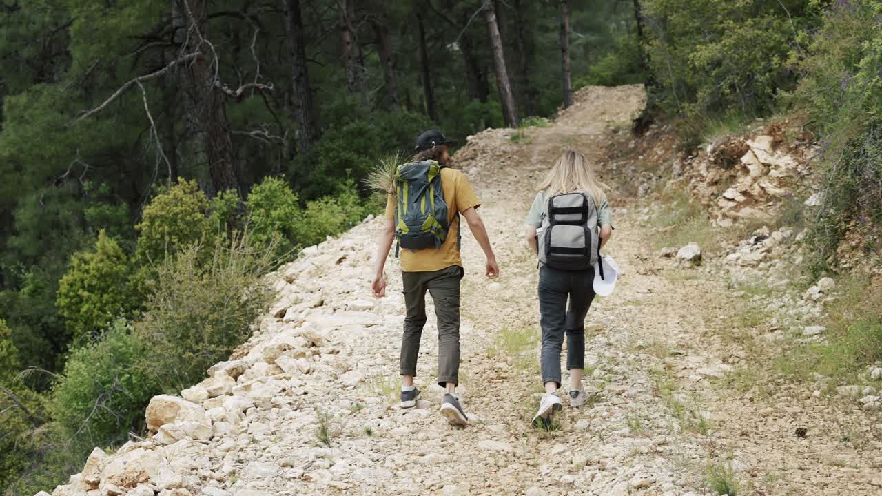 A happy couple backpackers walking by forest's rocky path, rear view