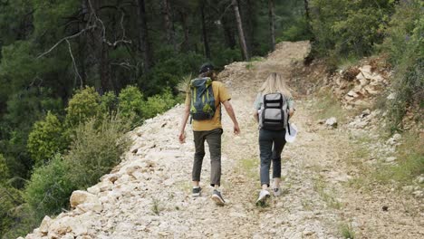 A-happy-couple-backpackers-walking-by-forest's-rocky-path,-rear-view