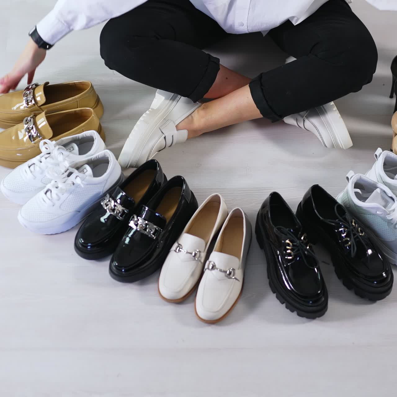 New pairs of modern footwear on the floor in front of sitting lady. Woman runs her hands over the diverse shoes choosing one black lacquered loafer. View from above