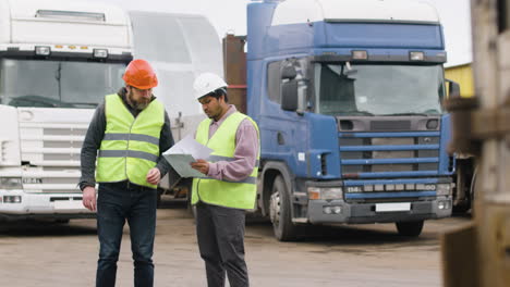 vista in lontananza di un capo e di un lavoratore che indossano giubbotti e caschi di sicurezza che organizzano una flotta di camion in un parco logistico mentre consultano un documento