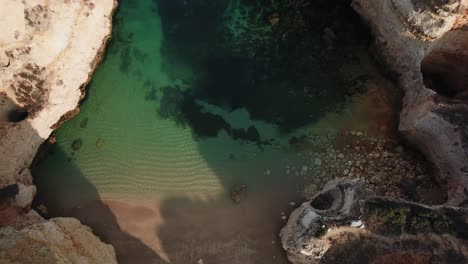 Rising-above-Algarve-beach-cove-as-clear-sea-water-washes-ashore,-Portugal,-birds-eye-aerial