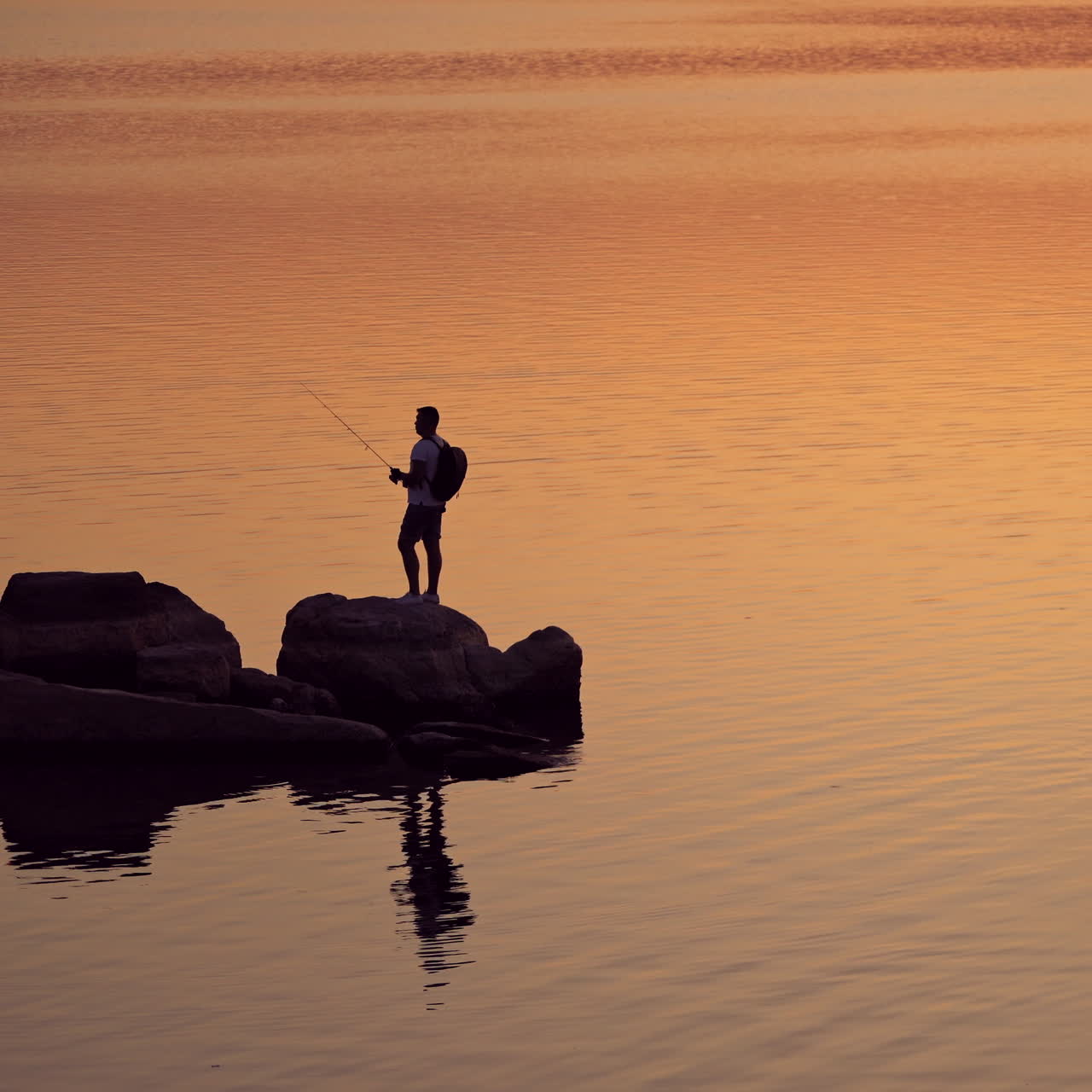 Silhouette of a fisherman with rod on the orange water background. Man with fishing rod is standing on a rock at a beautiful sunset in water.