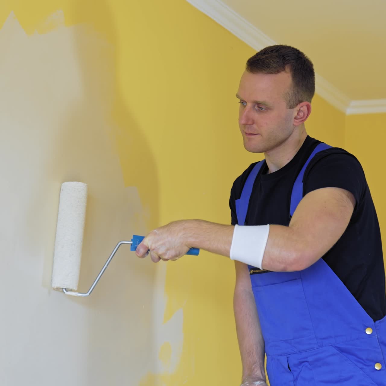 Professional painter paints the walls with roller brush. Worker man in blue overalls renovating and improving flat. Repair and decorating