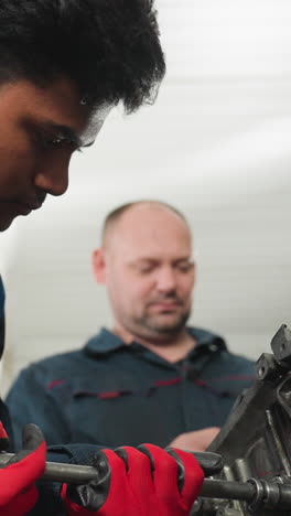 primer plano de un mecánico con pendiente magnético y guantes rojos soltando una nuez en un motor, observado atentamente por su colega en el fondo de un taller ocupado