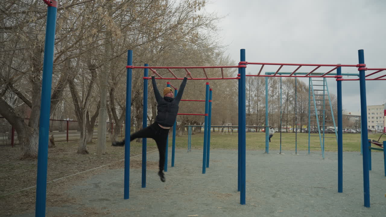 A coach leaps up to grab a red iron bar, then moves with controlled precision as he swings across it, in the background, a boy plays football on a field with park cars seen from a distance