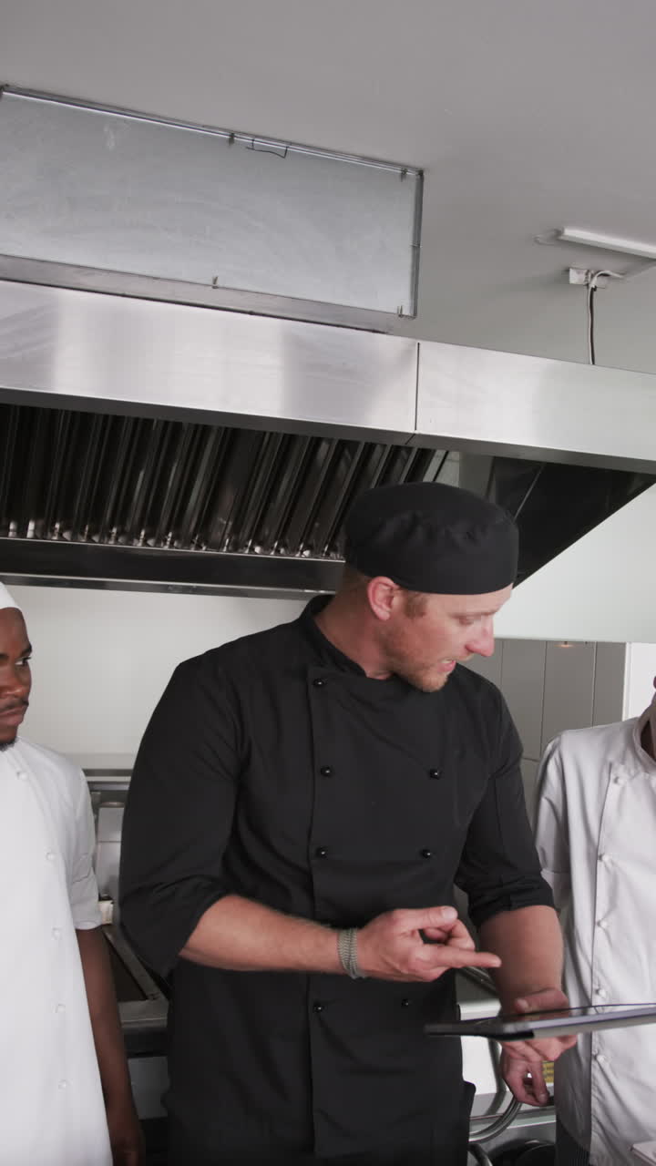 Focused diverse male chef instructing trainee male chefs in kitchen, slow motion, vertical