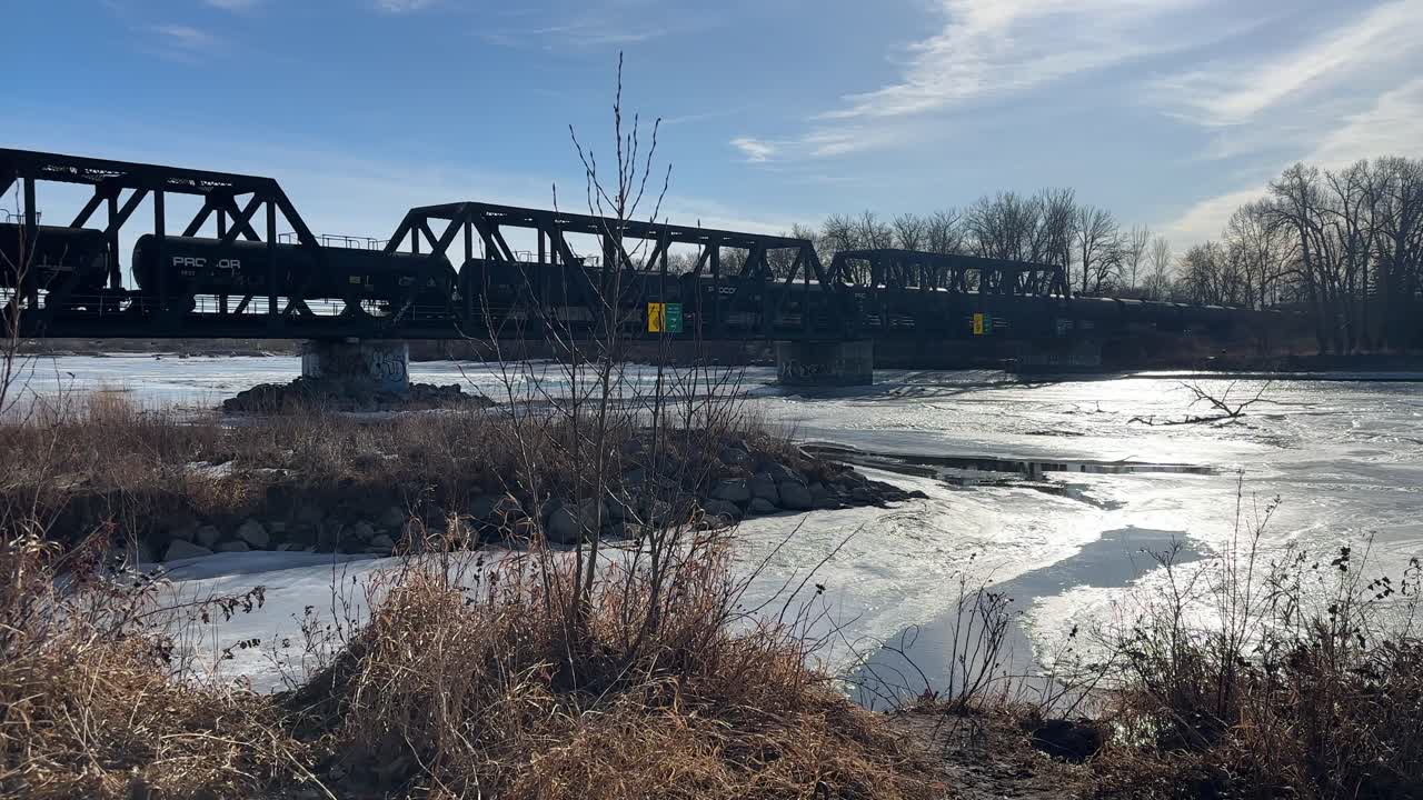 Cargo train tanks crossing bridge frozen river sunny day, Calgary, Alberta, Canada