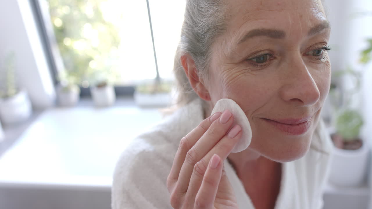 Applying skincare, mature woman using cotton pad on face in home bathroom
