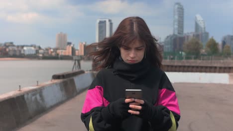 Mujer-Joven-Preocupada-Charlando-En-Su-Teléfono-Móvil-Mientras-Camina-Cerca-De-Un-Río-En-La-Ciudad-Al-Mediodía